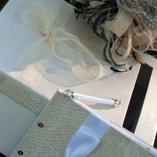 a set of custom python textured ecru card box, sign in book with silk ribbon, and Swarovski crystals encrusted sign in pen for a wedding displayed elegantly next to straw hat with ostrich feathers